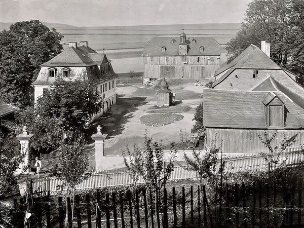 Historische Aufnahme: Blick über einen Lattenzaun auf einen Hof mit großen Gebäuden, dahinter Felder