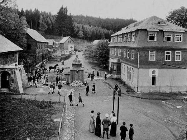 Historische Aufnahme: Menschen in langen Kleidern und Hüten auf der Dorfstraße vor großen Schieferhäusern