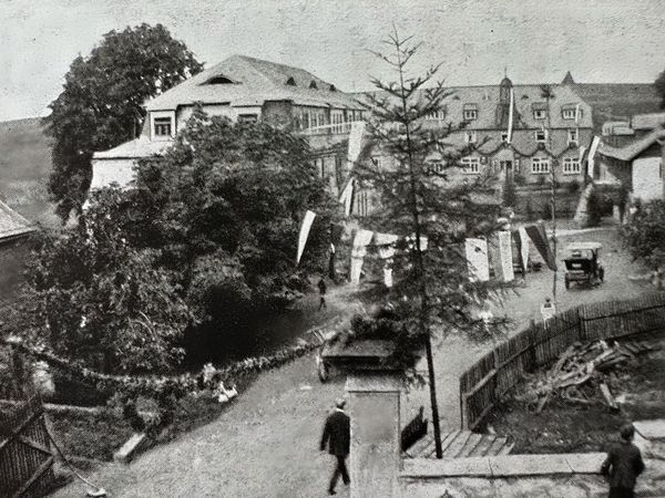 Historische Aufnahme: mit Fahnen und Girlanden geschmückte Dorfstraße mit Passanten und frühem Automobil