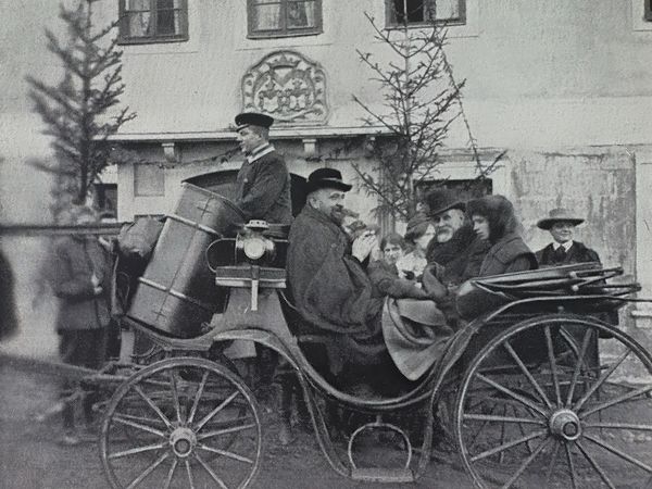 Historische Aufnahme: Fahrgäste in Mänteln und Hüten in einer offenen Kutsche vor einem Haus mit Wappen
