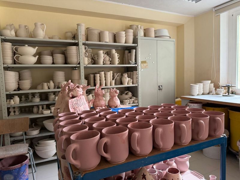 Wagen mit rosa Tassen und Katzenfiguren vor Regalen voller Rohware