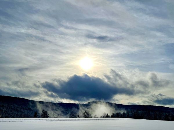 Wintersonne hinter Wolken über verschneiten Feldern und bewaldeten Hügeln mit Nebel