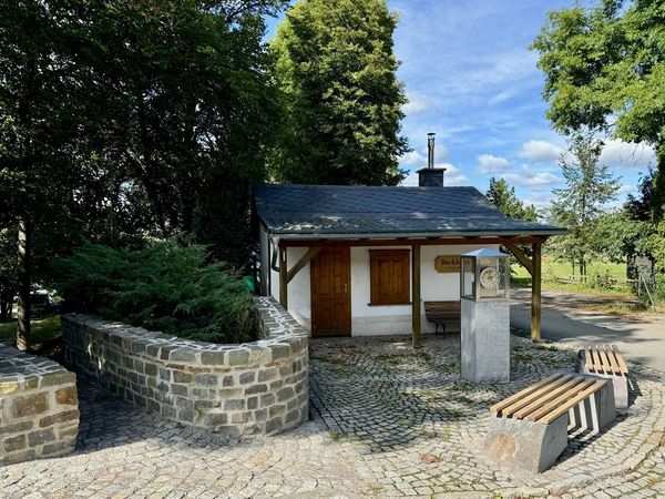 Backhaus mit geschwungener Natursteinmauer, Uhrensäule und Sitzbänken auf gepflastertem Platz